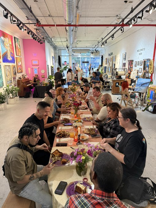 A long communal dining table filled with people enjoying a meal together in what appears to be an art gallery or creative space, with various artworks displayed on the walls around them.