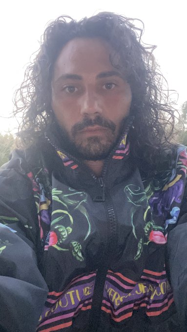 A close-up selfie of a man with curly dark hair and a beard, wearing a dark jacket over a brightly patterned shirt with green and pink designs.