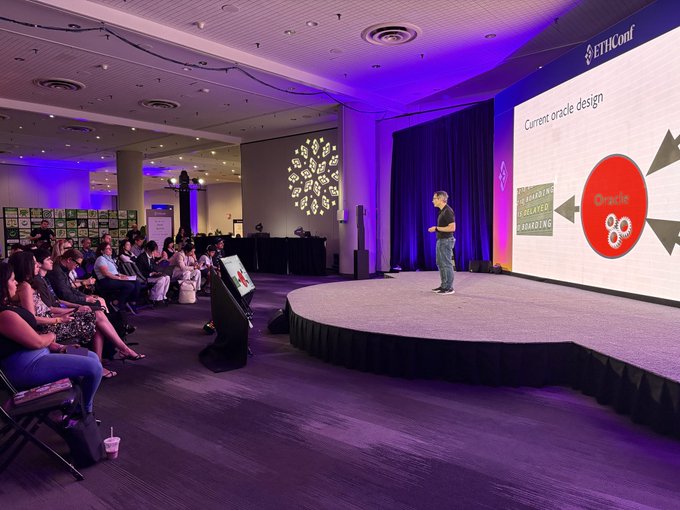 In the image, a man is standing on a stage giving a presentation while several people are sitting in chairs around him. The audience appears engaged as they listen to the speaker's lecture. A large screen behind the speaker displays information about the survey being presented by the speaker.