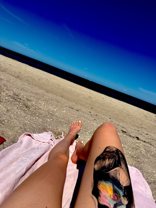 A person relaxes on a pink towel at the beach, with a colorful tattoo visible on their leg against a backdrop of sand and blue sky.
