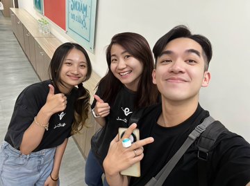 Two smiling women and a man pose for a selfie at the Virtuals Build & GenAI Fund workshop, with the woman in the middle proudly displaying the "Virtuals" key she received for vibe coding. A "Diva Karaoke" sign is visible in the background hallway.