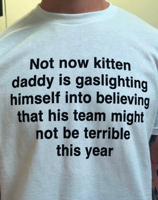 A person wearing a white t-shirt with the humorous text: 'Not now kitten daddy is gaslighting himself into believing that his team might not be terrible this year'.