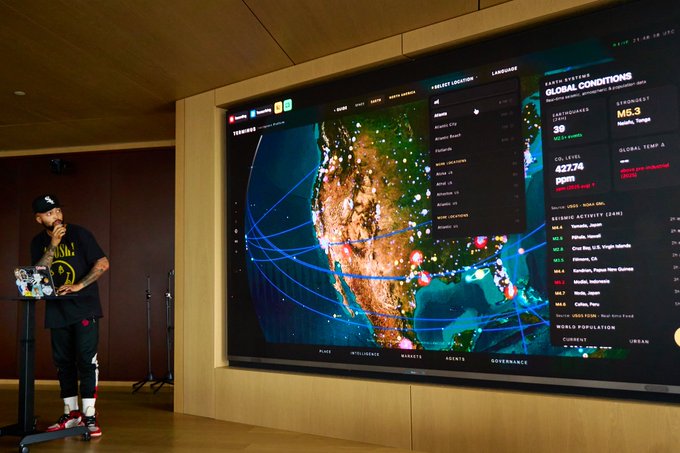 The image shows a man standing behind a large screen displaying a map of the world with various data points and information about global issues such as climate change, population growth, and more. The man appears to be giving a speech or presentation related to these topics. In addition to the main subject, there is also an image of a person holding a book in the scene.