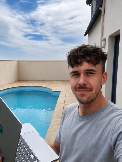 A young man with curly dark hair and a light blue t-shirt taking a selfie outdoors by a swimming pool while holding an Apple MacBook.