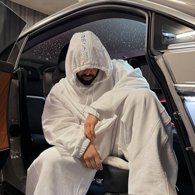 A bearded man sits in a luxury vehicle (appears to be a Rolls Royce) wearing a white hooded sweatshirt with colorful embroidered text, with a starlight headliner visible above him.