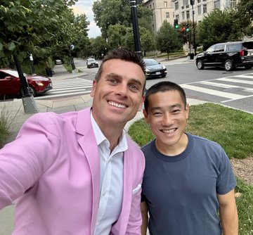 Two smiling men take a selfie outdoors on a city street, one wearing a pink blazer and the other in a blue t-shirt.