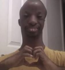 A black man wearing a yellow shirt, smiling and making a heart shape with his fingers in front of his chest.