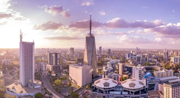 An aerial view of a city skyline at sunset, featuring a tall distinctive tower with a pointed spire, modern high-rise buildings, and uniquely shaped domed structures in the foreground.