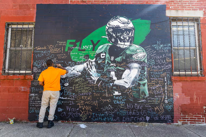 A man in an orange shirt stands before a large street mural dedicated to 'F1 MVP' (Jalen Hurts of the Philadelphia Eagles), covered in fan messages and signatures.