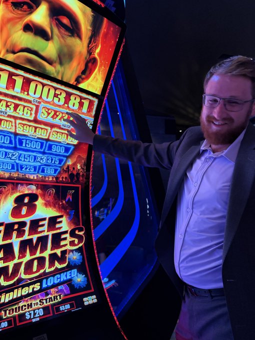 A smiling bearded man in glasses, a white shirt, and a dark blazer stands next to a brightly lit '8 Free Games Won' slot machine, pointing at a winning line showing 11,003.81.