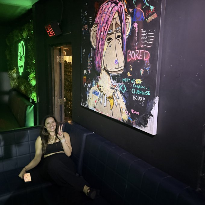 A smiling woman in a black outfit sits on a blue leather bench in a dimly lit venue, posing with a peace sign beneath a colorful painting of a Bored Ape with pink dreadlocks and the word 'BORED'.
