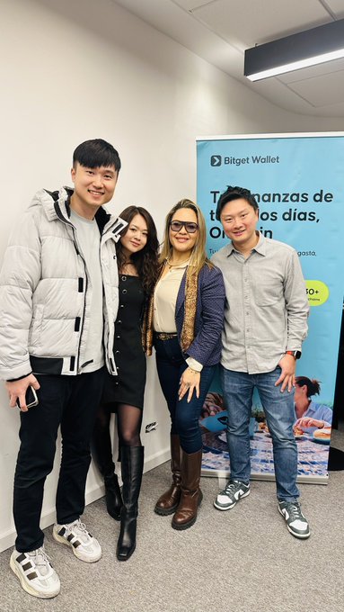 The image shows four people posing together at what appears to be an office visit, standing in front of a "Bitget Wallet" banner featuring Spanish text that reads "Tus finanzas de... tus días" along with a "10%" promotion. The group is dressed in business-casual attire, with one woman wearing glasses and a blazer, another in a black dress and boots, and two men in jackets and jeans, all smiling warmly for the photo—reflecting the post's theme of a special meeting with the Bitget Wallet team visiting from Asia.