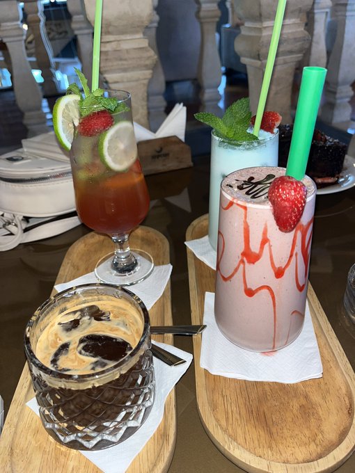 A wooden serving board holds an assortment of colorful beverages, including a red drink with a green straw, a tall pink milkshake topped with a strawberry, and a glass containing a creamy dessert with coffee beans.