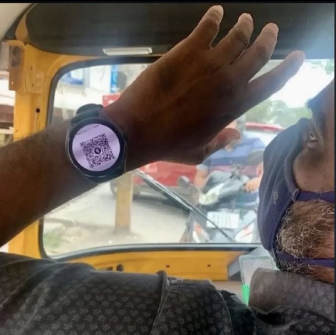A person displays a smartwatch on their wrist showing a QR code screen, inside what appears to be an auto-rickshaw with a driver visible in the background.