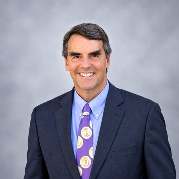 Tim Draper, a well-known Bitcoin advocate and venture capitalist, smiles warmly in a professional headshot wearing a dark suit, light blue shirt, and a distinctive purple tie featuring Bitcoin "₿" symbols. The tie's prominent Bitcoin logo ties directly to his recent comments urging major corporations like Apple and Meta to add Bitcoin to their balance sheets.