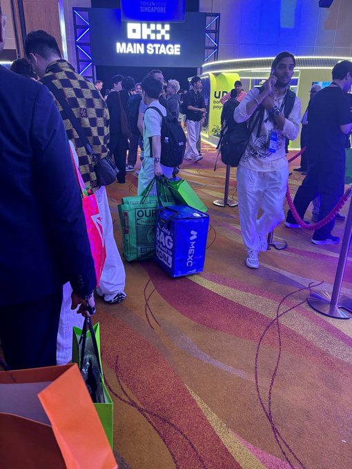 People walk through a convention venue near a 'TOKEN 2049 MAIN STAGE' sign, carrying branded shopping bags including one reading 'MEXC'.