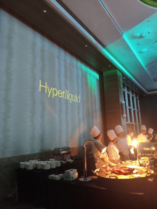 Chefs in white uniforms and tall hats work at a buffet station in a modern venue where the word 'Hyperliquid' is projected in yellow on a large screen.