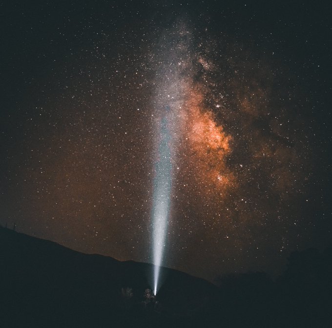 A stunning astrophotography image capturing the Milky Way galaxy's colorful core stretching across a starry night sky. In the foreground, the dark silhouettes of mountains frame a person standing and shining a bright flashlight beam straight up toward the galaxy.