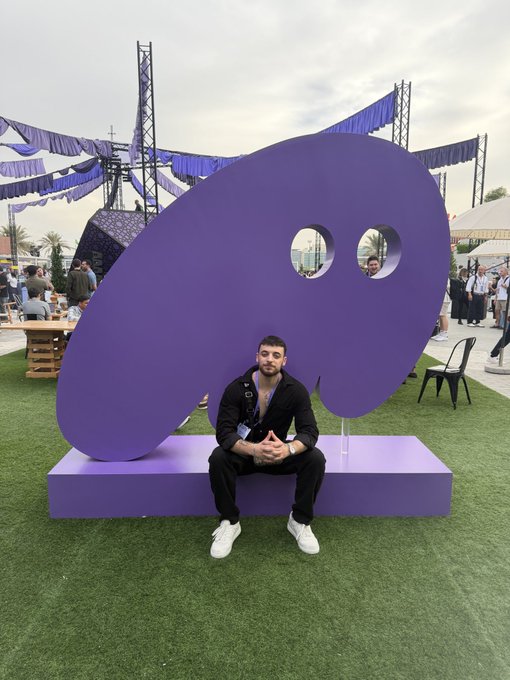 A man sits on a purple bench in front of a large purple artist's palette sculpture at what appears to be an outdoor art festival or event. The "GM" (Good Morning) post likely refers to a cheerful morning greeting shared from this creative, visually striking setting.