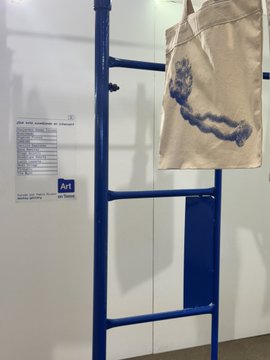 A beige canvas tote bag with a blue abstract print hangs on a blue metal ladder display at the Tucumán Contemporary Art Fair (1st edition), alongside an information placard marked "A4" listing the participating artists.
