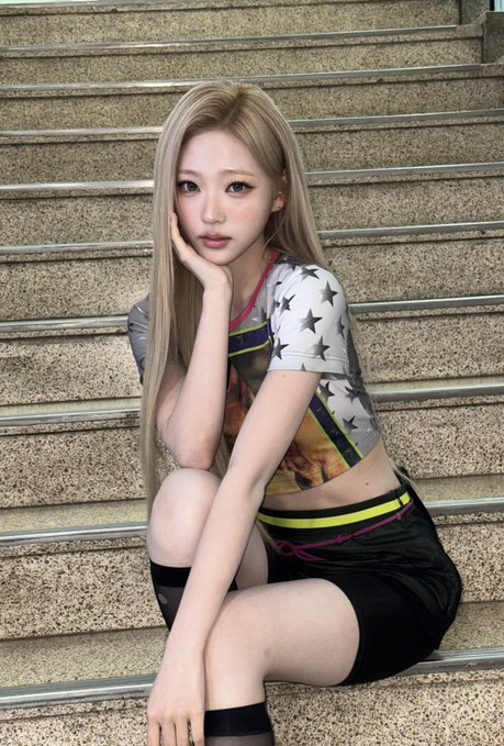 Young woman with long blonde hair sitting on granite stairs, wearing a star-patterned cropped top, black shorts with yellow trim, sheer black socks, and small heart makeup under her eye.