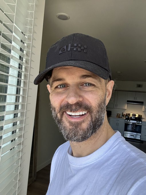A bearded man wearing a black "d" branded cap and white t-shirt smiles for a selfie in his modern kitchen. The relaxed, upbeat vibe of the shot perfectly matches the cheerful "Gm! Happy Sunday degens" greeting.