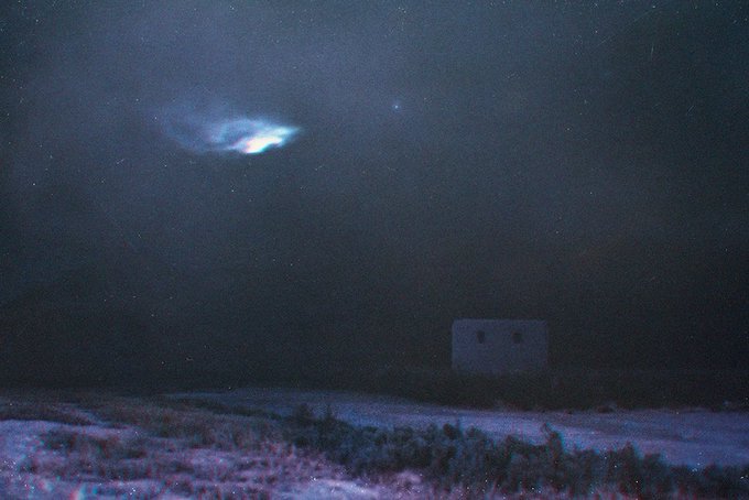 A grainy nighttime photograph showing a glowing blue UFO-like light in the sky above a dark desert landscape with a small building in the distance.