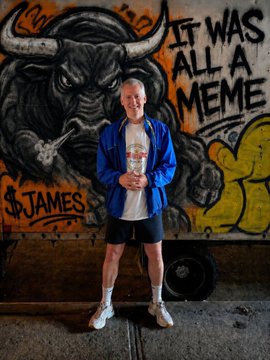 A man in a blue jacket and shorts stands smiling in front of a graffiti mural featuring a bull, with the text 'IT WAS ALL A MEME' and '$JAMES' visible on the wall.