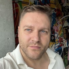 A selfie of a man with short brown hair and blue eyes wearing a white shirt, standing in front of a graffiti-covered wall.