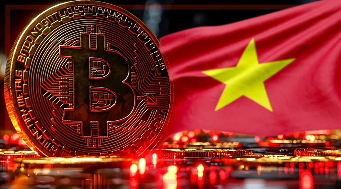 A close-up of a gold Bitcoin coin with inscriptions including 'BITCOIN DIGITAL DECENTRALIZED PEER TO PEER' standing upright, with the red Vietnamese flag featuring its yellow star blurred in the background.