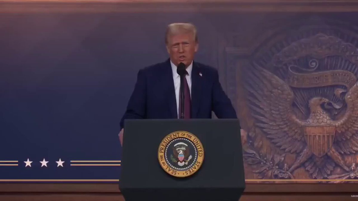 The image features a man standing at a podium, giving a speech or presentation while wearing a suit and tie. The setting appears to be an auditorium with a stage designed like a wing. The main focus of the image is the man speaking from behind the podium.
