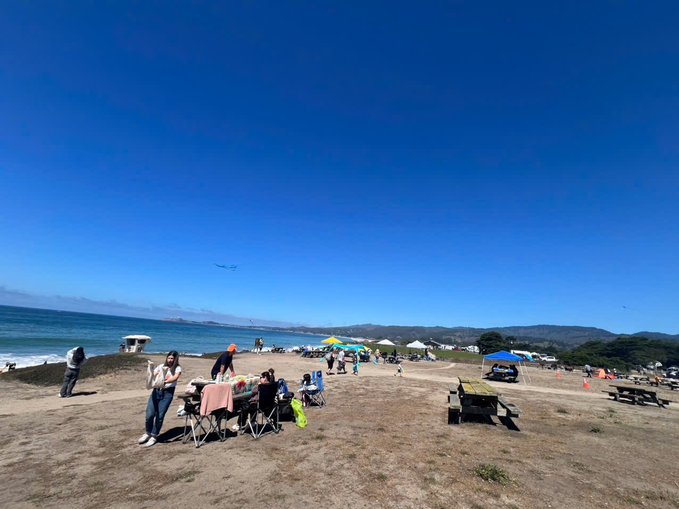 A sunny beach scene with people sitting on folding chairs and under canopies near the shoreline, with mountains visible in the distance and an airplane flying overhead.