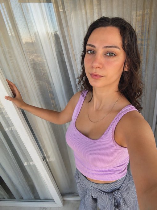 A mirror selfie of a woman with wet dark hair wearing a light purple ribbed tank top and grey pants, standing by a window with sheer white curtains.