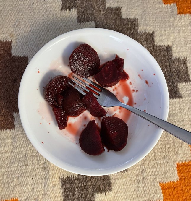 A top-down view shows a white bowl containing sliced, dark red beets with a metal fork resting in the juice, placed on a patterned rug.