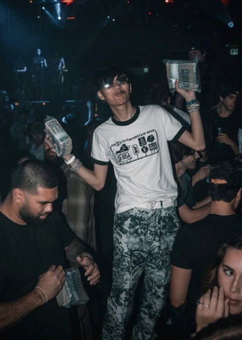 A young man in a graphic t-shirt and floral pants stands in a crowded club holding two clear boxes, surrounded by other partygoers in low light.