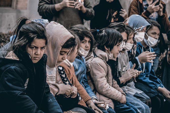 A group of children, some wearing face masks, sit closely together with somber expressions, embodying the post's poignant theme of innocence overshadowed by difficult circumstances. The visible "UNITED" text on one child's jacket subtly reinforces the message of solidarity with those born into hardship.