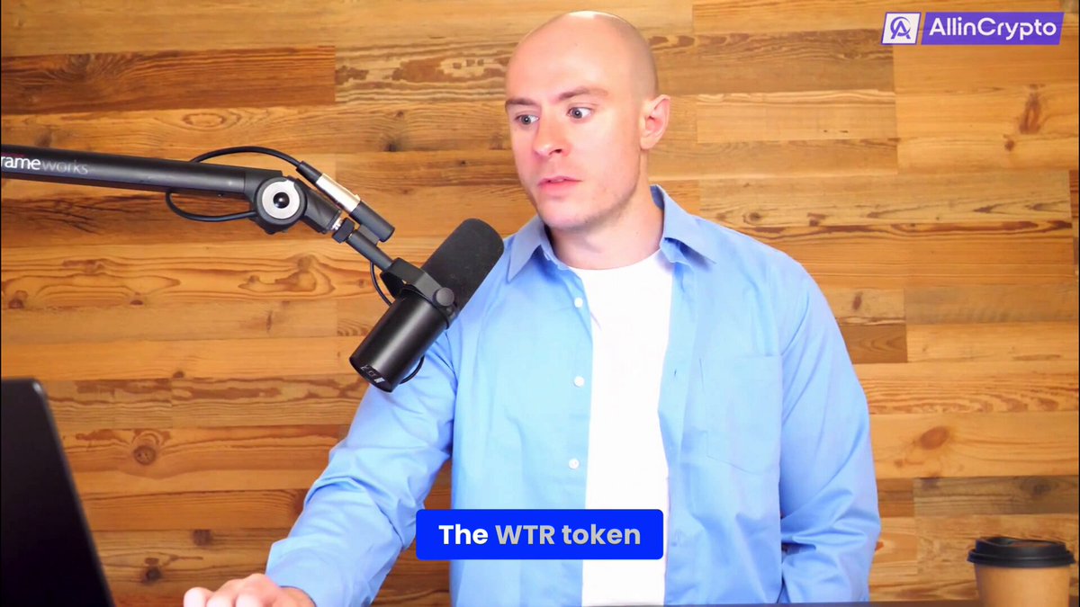 The image shows a man wearing a blue shirt standing behind a microphone at a desk with a laptop computer. The background features wooden panels, giving an office setting. There are also two cups visible in the scene - one near the right edge of the frame and another closer to the center.