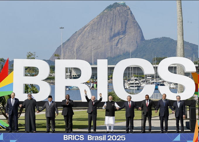 Leaders stand hand-in-hand beneath large white "BRICS" letters with "BRICS Brasil 2025" displayed below, set against Rio de Janeiro's iconic Sugarloaf Mountain, capturing the diplomatic unity of the summit. The image reinforces the post's message by visually representing Brazil's commitment to the BRICS alliance, de-dollarization efforts, and an independent foreign policy—highlighting that opposing these stances means going against Brazil's national interests.