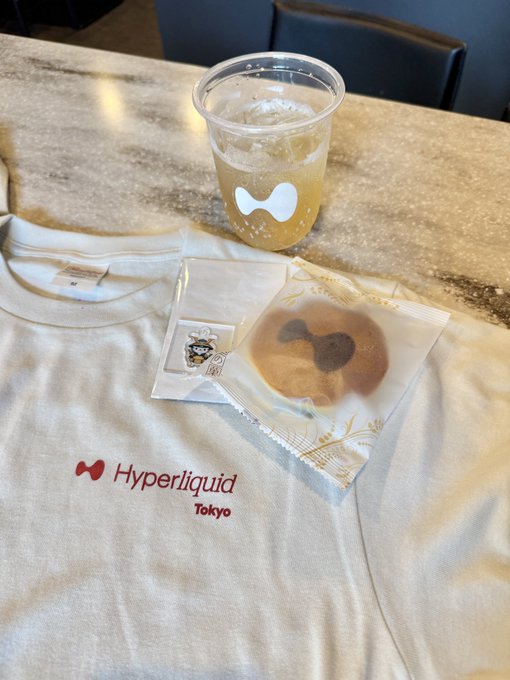 A "Hyperliquid Tokyo" t-shirt laid out on a table alongside an iced drink and a packaged snack featuring the Hyperliquid logo, likely given as merchandise at the Hyperliquid Japan gathering event.