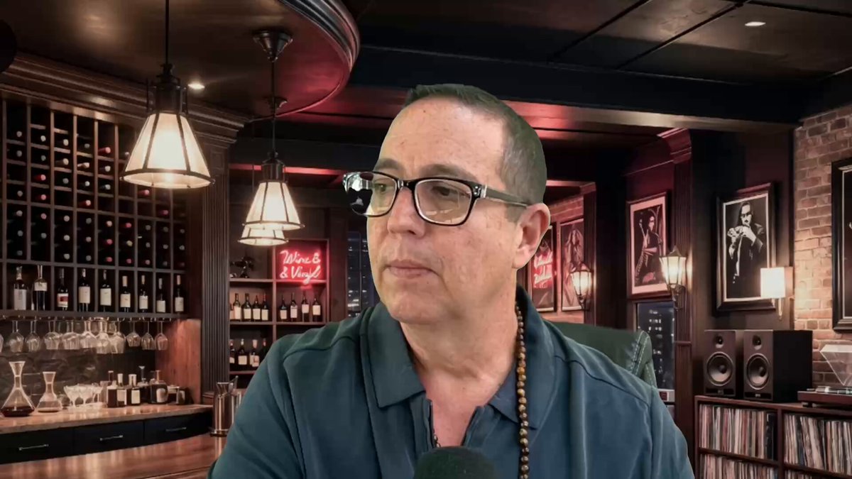 The image features a man wearing glasses, sitting at a bar with several bottles of wine behind him. He appears to be engaged in conversation or observing something off-camera. The background includes a dining table surrounded by chairs, suggesting that this could be an upscale restaurant or bar setting. There is also a neon sign visible in the scene, which might indicate the name of the establishment or a special event taking place at the venue.