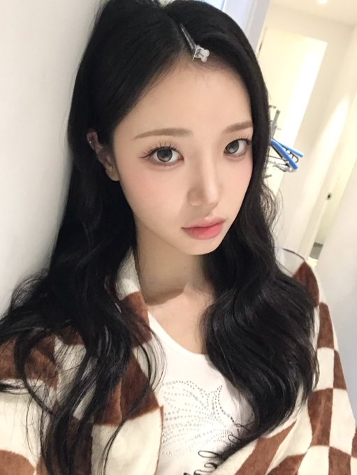 Close-up selfie of a young woman with long dark hair wearing a white top and a brown and white checkered cardigan, with a small hair clip visible in her bangs.