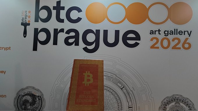 In the image, there's a poster for an art gallery event called "BTC Prague 2016". The poster features a red banner with white text that says "BTC Prague 2016" and "The Art Gallery". There are also two circles on the poster - one orange circle labeled "BTC" and another orange circle labeled "PRague". A book is placed in front of the poster, which has an interesting design. It's a leather-bound book with a gold cover featuring a red Bitcoin symbol. The book appears to be made by @TheLeatherMint.