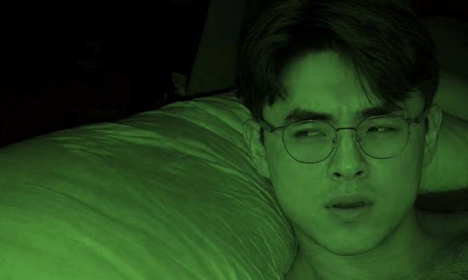 A close-up portrait of a man with glasses lying in bed, lit by an eerie green light.