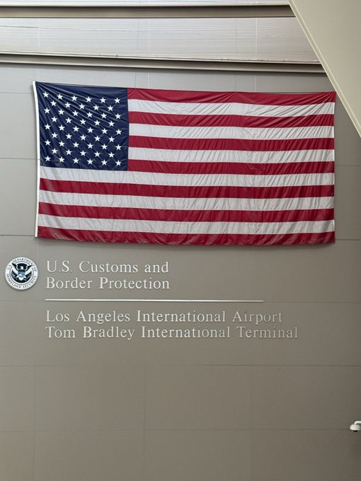 A large American flag mounted on a wall above signage reading 'U.S. Customs and Border Protection, Los Angeles International Airport, Tom Bradley International Terminal.'