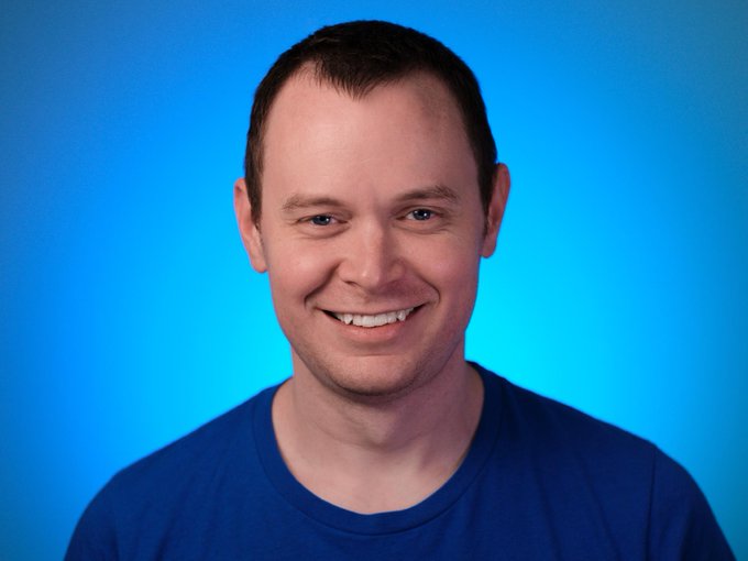 The image features a man wearing a blue shirt with a big smile on his face as he looks directly into the camera. The background of the photo is a gradient of blue that appears to be a light blue sky, which contrasts nicely with the man's bright smile and adds depth to the image. There are no other objects or people in the picture, making it a simple yet striking composition.
