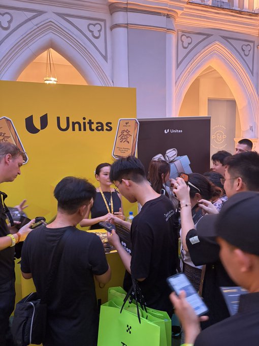 Crowd of people gathered at a yellow 'Unitas' branded booth, with attendees looking at phones and receiving items, set against arched ceilings in what appears to be a gothic-style venue.
