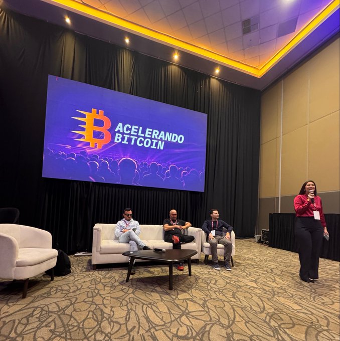 A photograph of a panel discussion at an event titled 'ACELERANDO BITCOIN,' featuring three men seated on a white sofa and a woman in a red shirt speaking into a microphone.