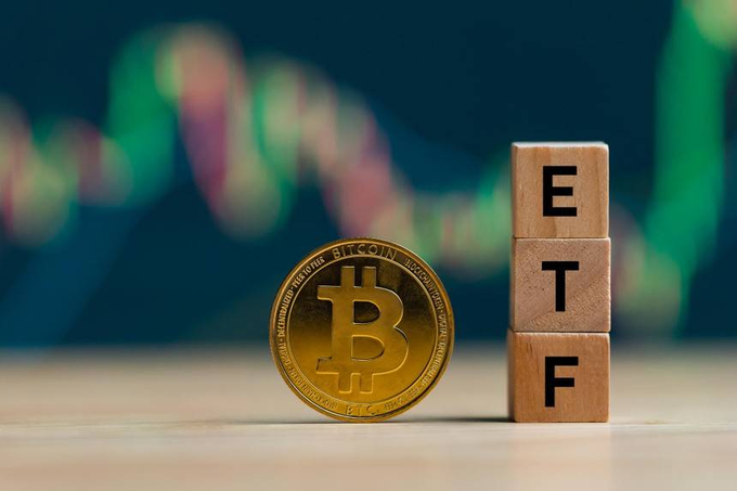 A physical Bitcoin token stands next to wooden blocks spelling 'ETF' with a blurred stock chart in the background.