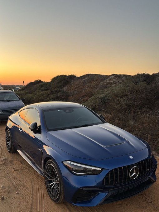 A blue Mercedes-AMG coupe parked on a sandy area with scrub brush and a sunset sky in the background.