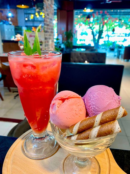 A red iced drink garnished with pineapple leaves sits beside a dessert of strawberry and purple ice cream scoops with wafer rolls, displayed in a cafe setting.